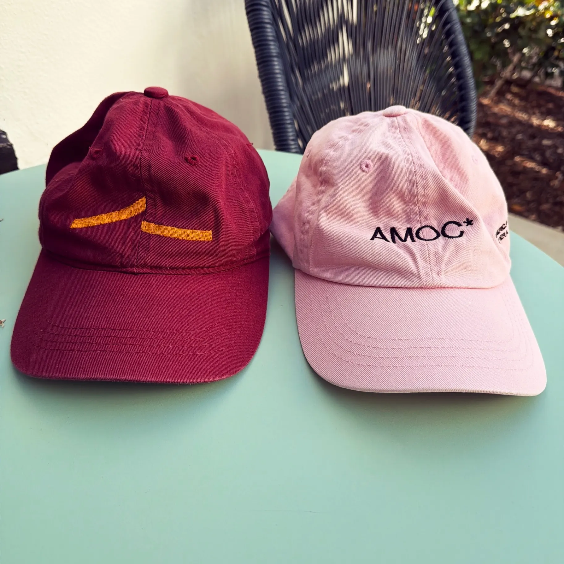 Two embroidered baseball caps on a mint-green table — a burgundy one with a gold X and a pink AMOC* cap from the American Modern Opera Company