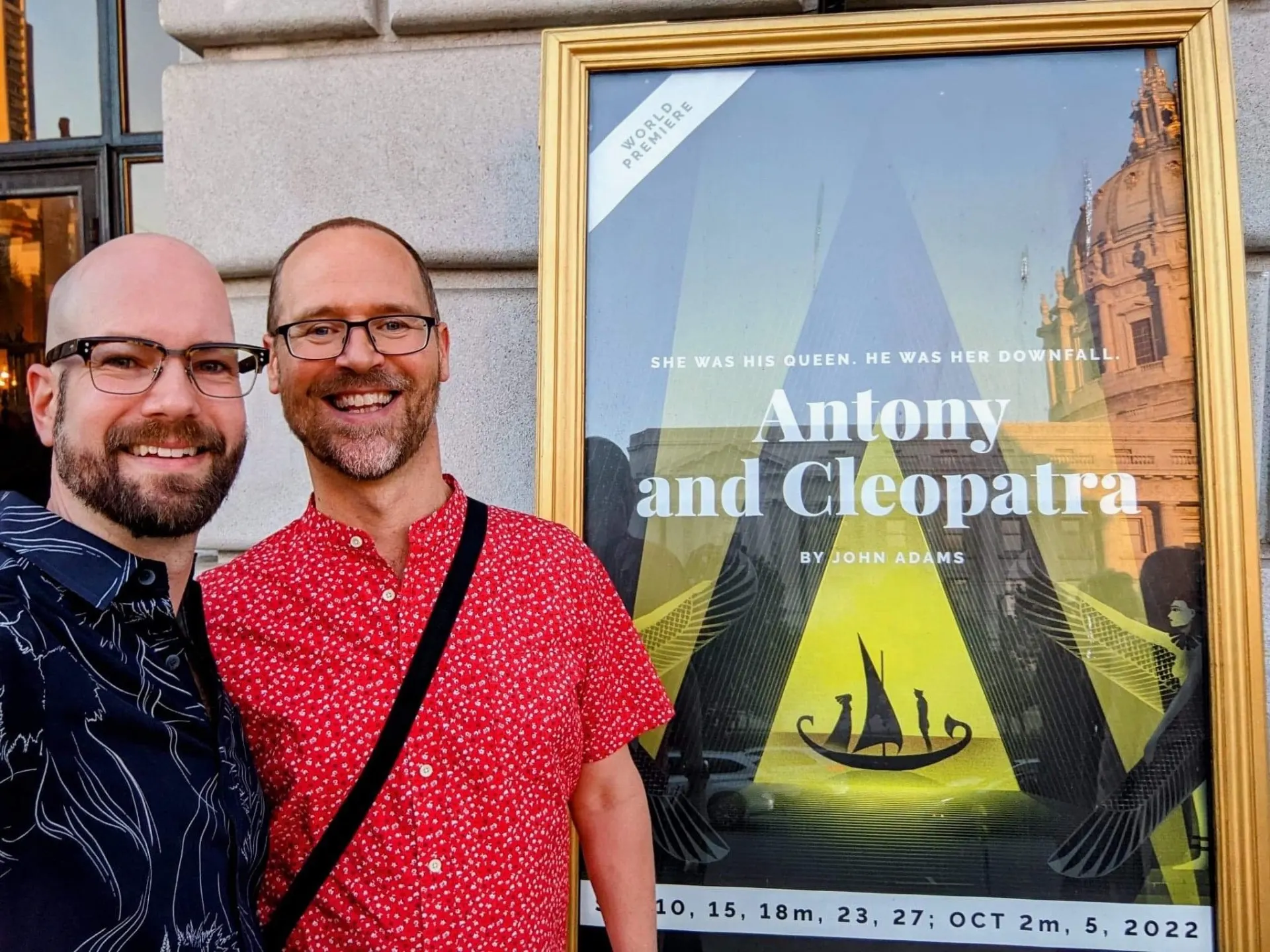 Two friends smiling in front of the Antony and Cleopatra world premiere poster at San Francisco Opera
