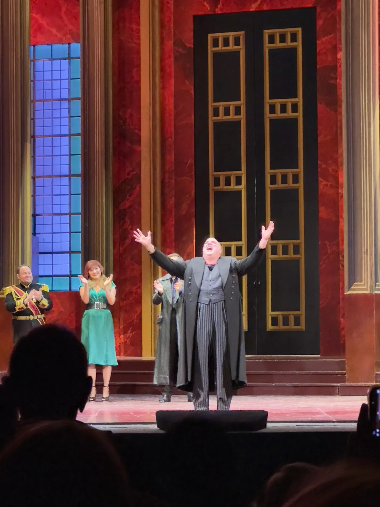 Baritone Michael Chioldi at curtain call with arms thrown wide, red marble set with tall black-and-gilt doors behind, colleagues applauding