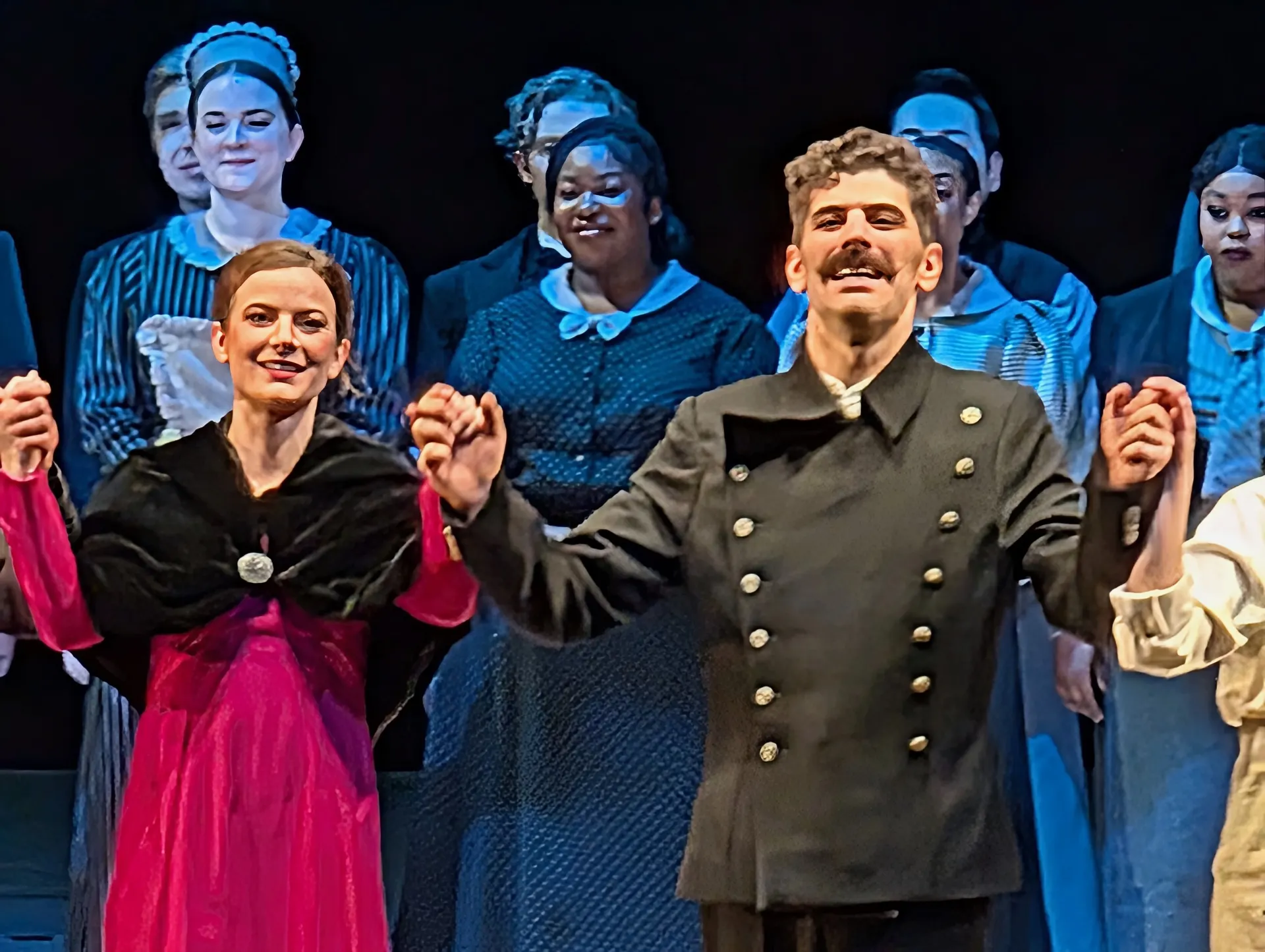 Curtain call principals of Smetana's Dalibor — a woman in a magenta gown and black cape beside the title character in a brass-buttoned military coat, chorus in 19th-century dress projected behind