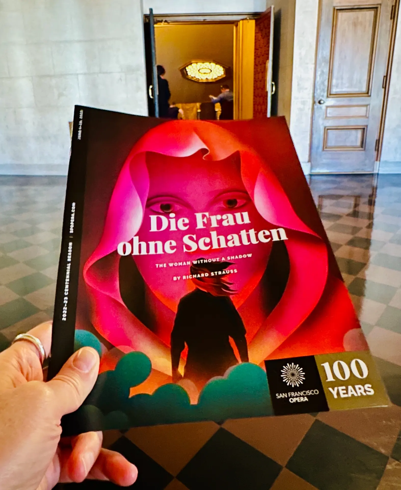 Die Frau ohne Schatten centennial program held in a marble lobby, with a glowing doorway and chandelier beyond