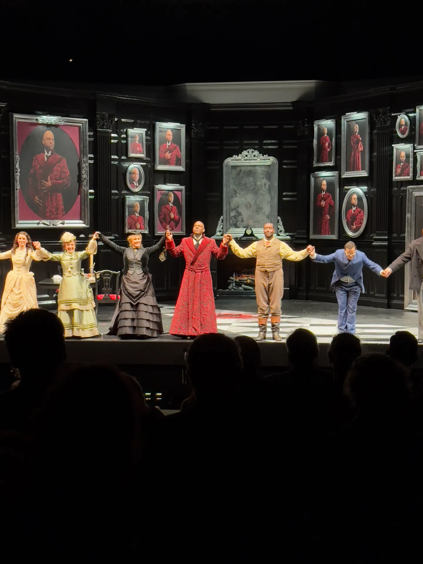 Full company of The Picture of Dorian Gray taking a bow on a black-paneled set hung with multiple framed portraits of the title character in a red brocade robe