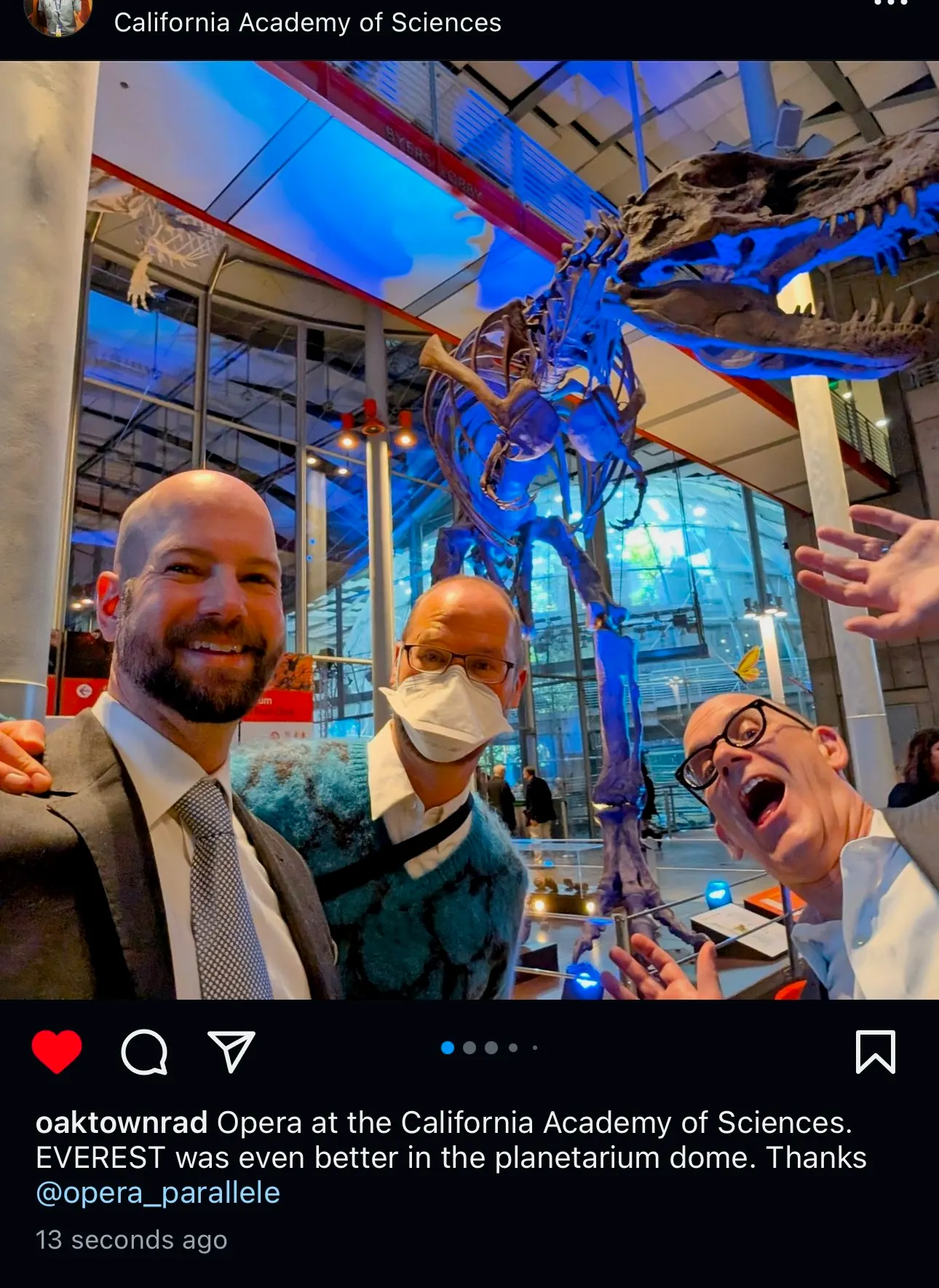Three-person selfie in the California Academy of Sciences atrium beneath the T. rex skeleton lit blue, one in mask and turtleneck, another in a suit, a third laughing in glasses
