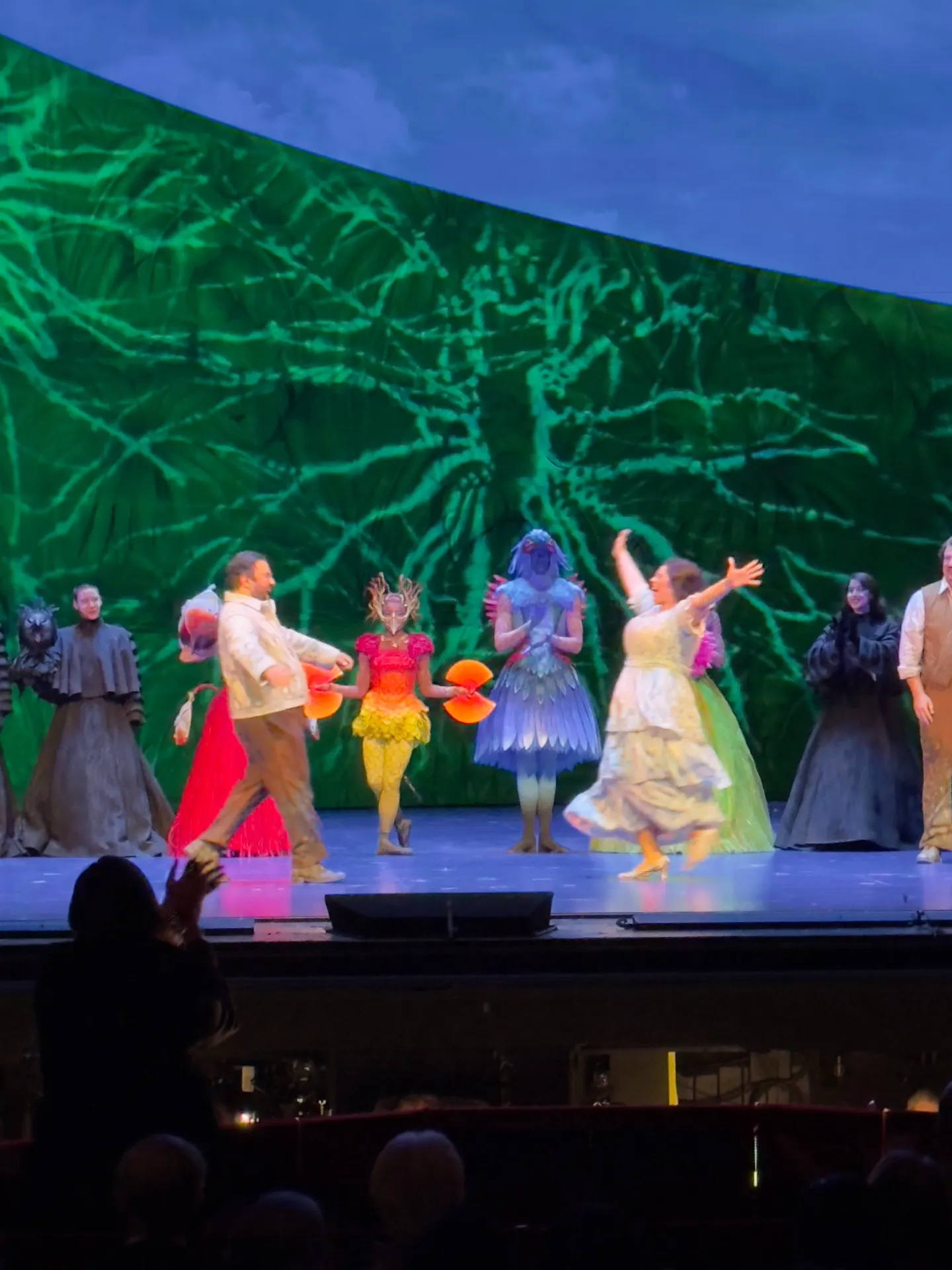 Florencia en el Amazonas curtain call — principals in jewel-toned costumes (red fringe, butterfly, peacock-feathered river spirit) bowing against a green jungle cyclorama