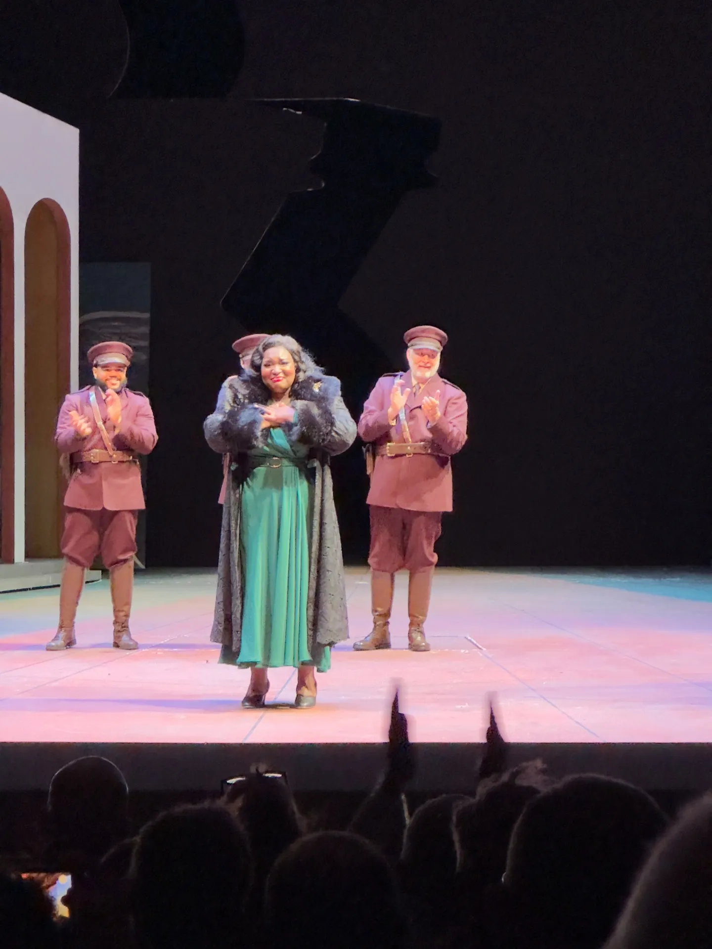 Singer in a green dress and dark coat taking a bow with uniformed castmates applauding behind her
