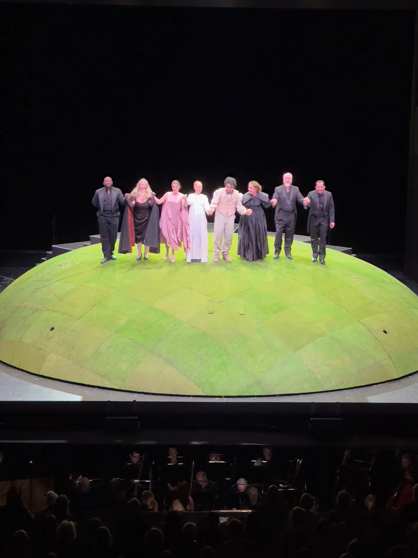 Curtain call on a giant green domed set with the cast lined hand-in-hand above the orchestra pit