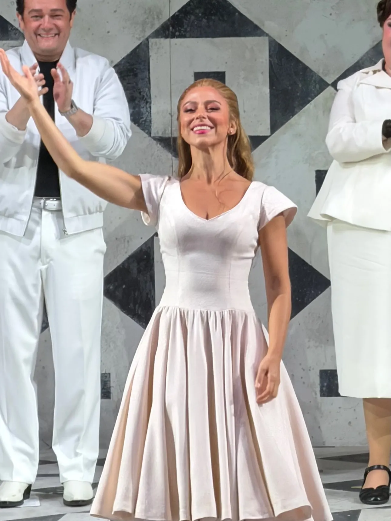 Dancer in a pale blush dress taking her bow at the Innocence curtain call, principals in white applauding beside her on a black-and-white diamond-tiled set