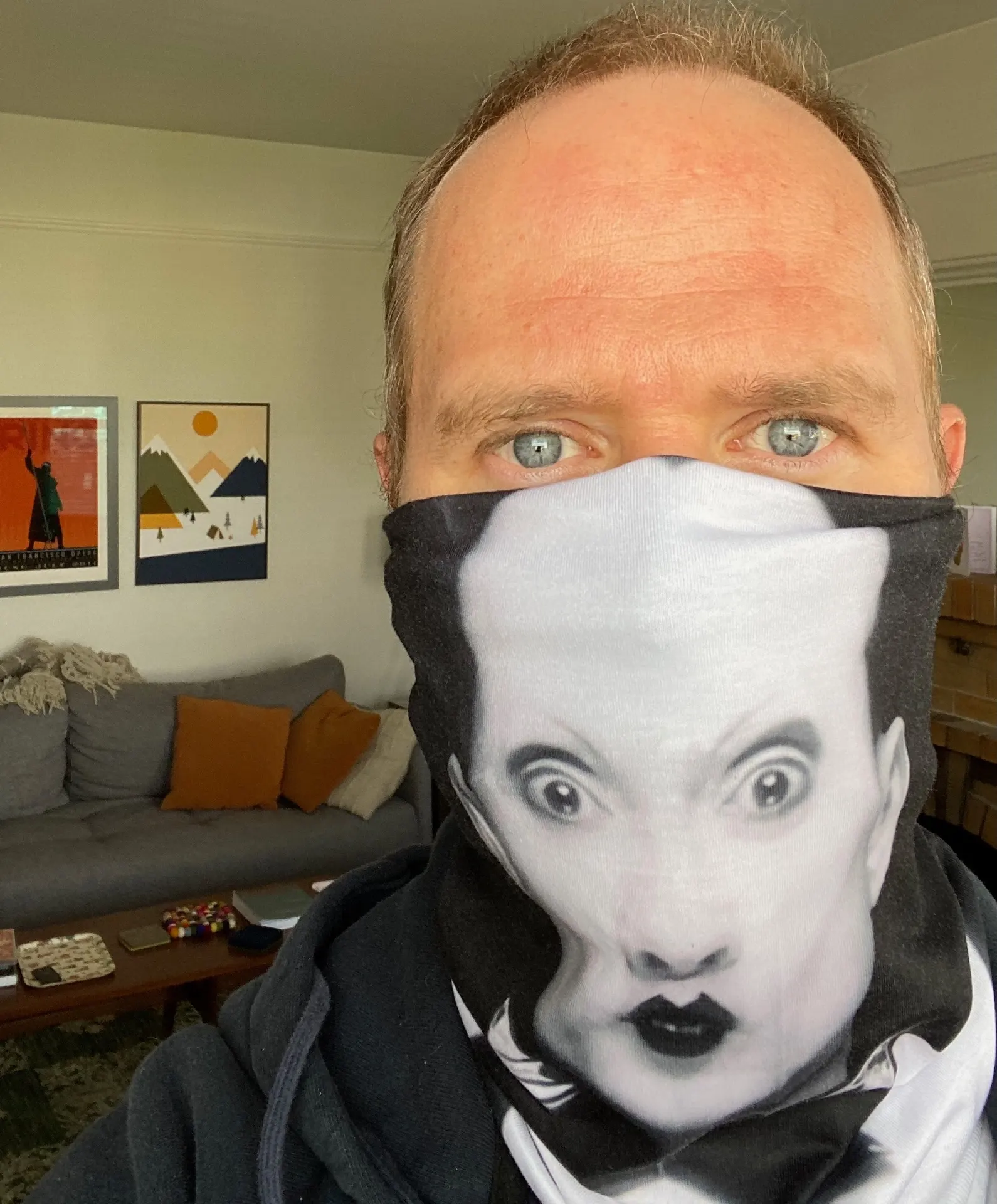 Selfie wearing a black-and-white face covering printed with Klaus Nomi's iconic made-up face, in a living room with an SF Opera poster on the wall
