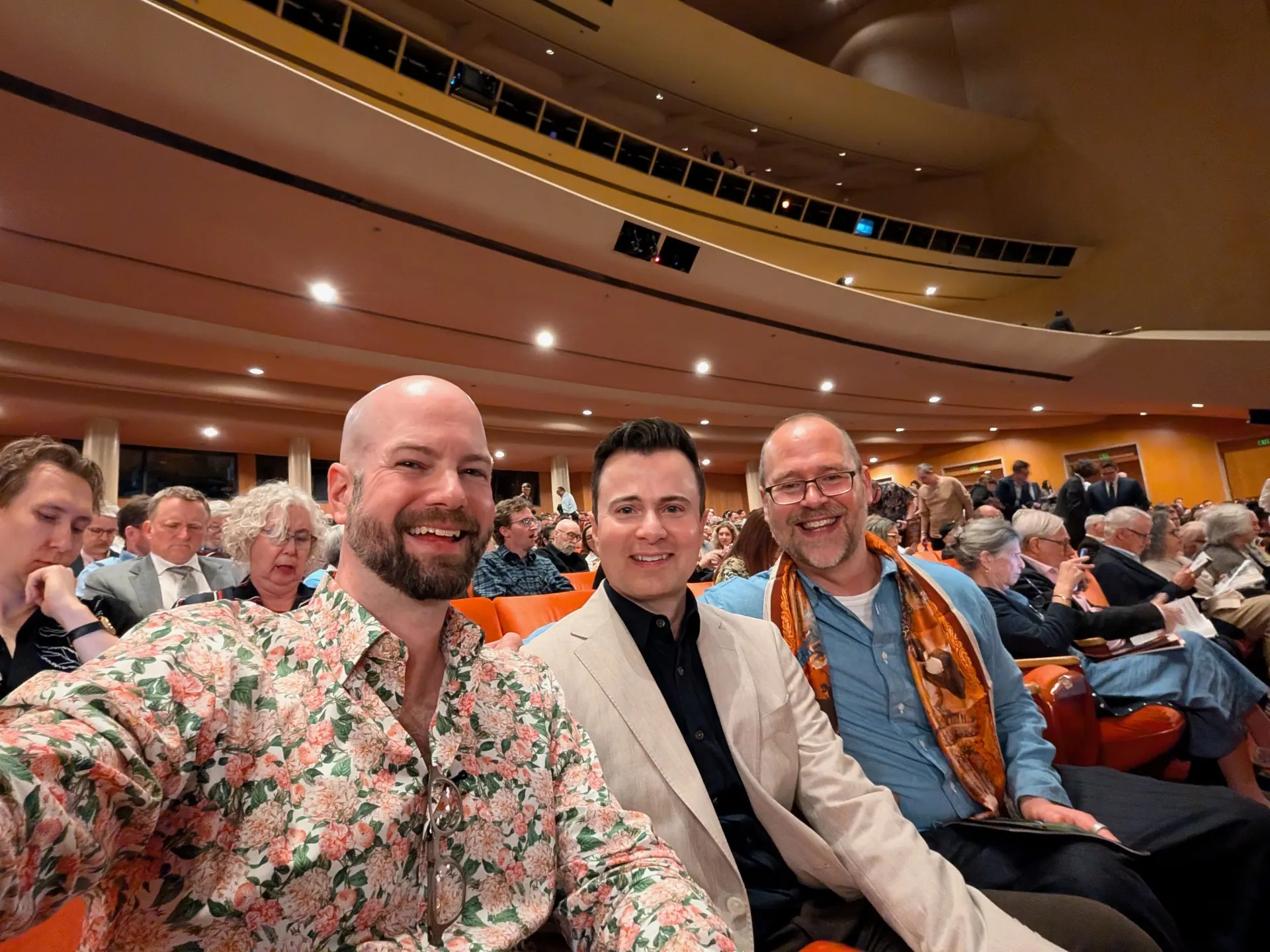 Three friends taking a seated selfie inside the Dorothy Chandler Pavilion before the performance, surrounded by rows of orange seats and the audience settling in