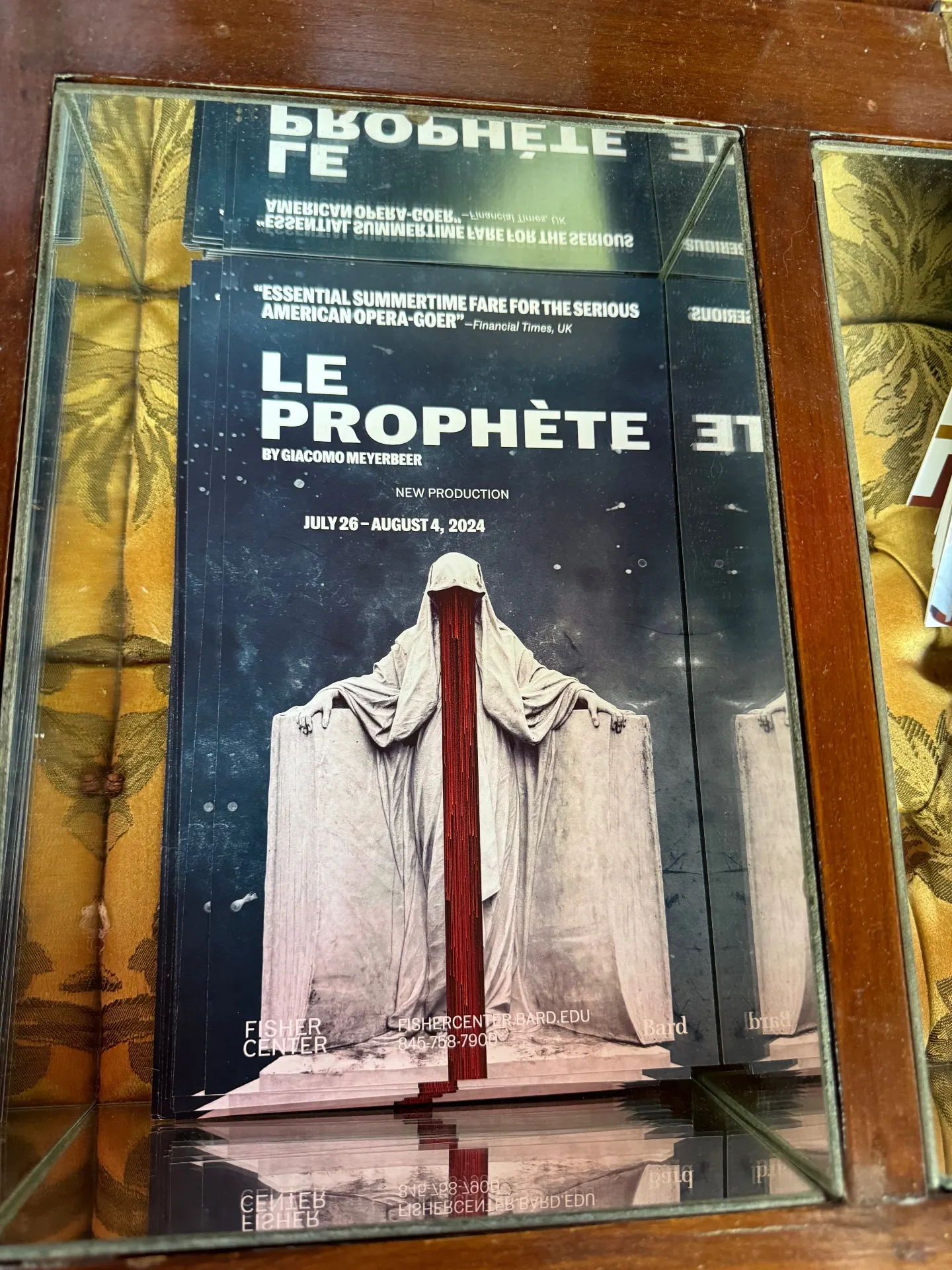 Stacked Le Prophète postcards in a mirrored lobby display at Bard's Fisher Center, white-marble shrouded figure with a red veil
