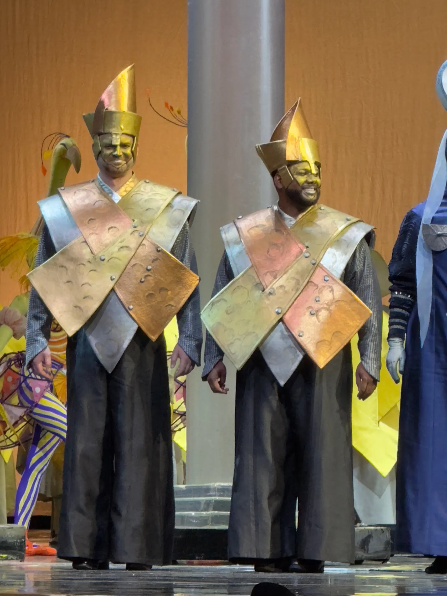 Two men in tall gold helmets and crossed bronze armor plates over chainmail, standing on stage in front of an orange backdrop with painted yellow flora