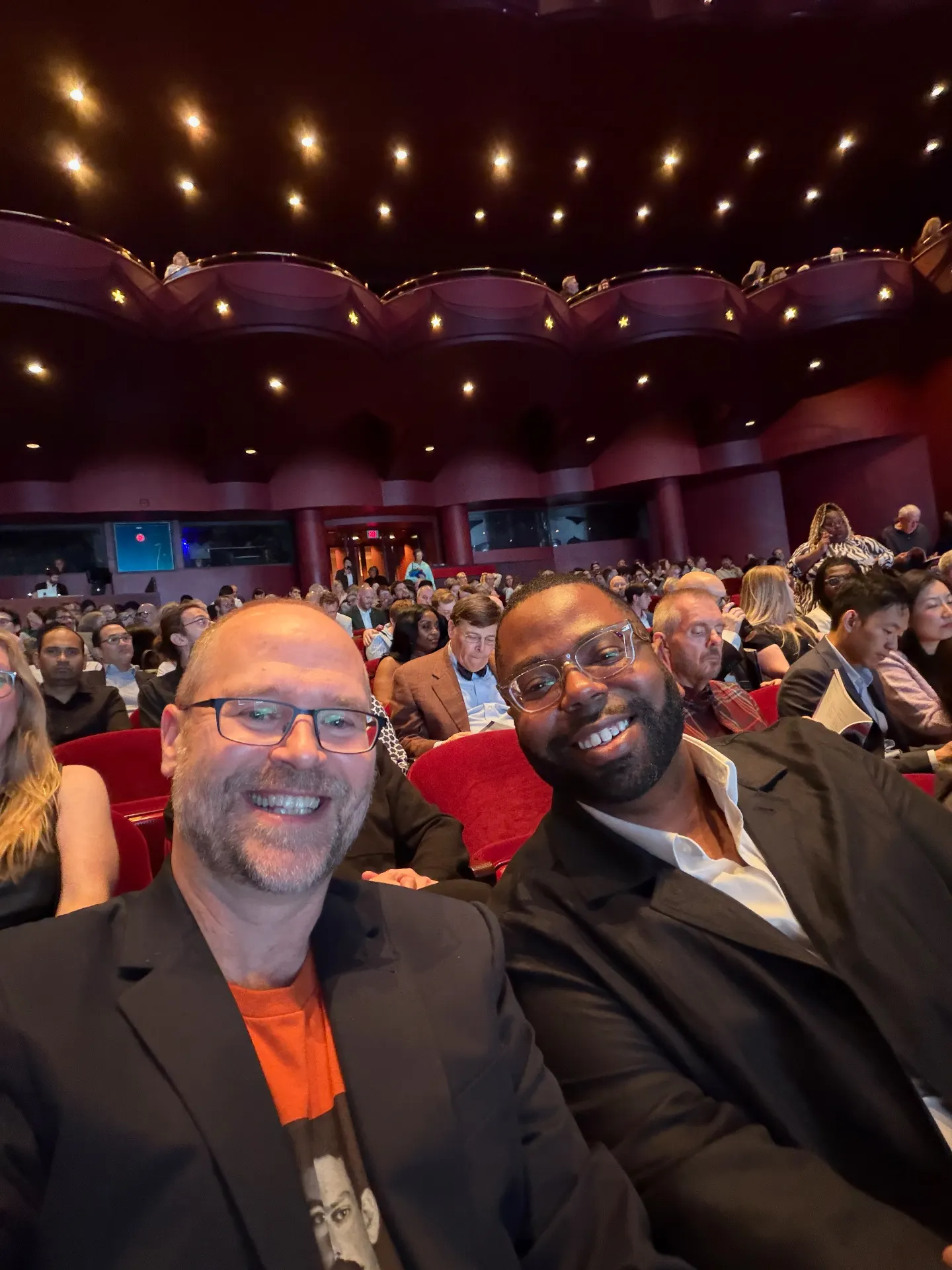 Two friends in dark blazers grinning for a selfie inside the Wortham Center, red seats and starlit ceiling behind them, audience filing in