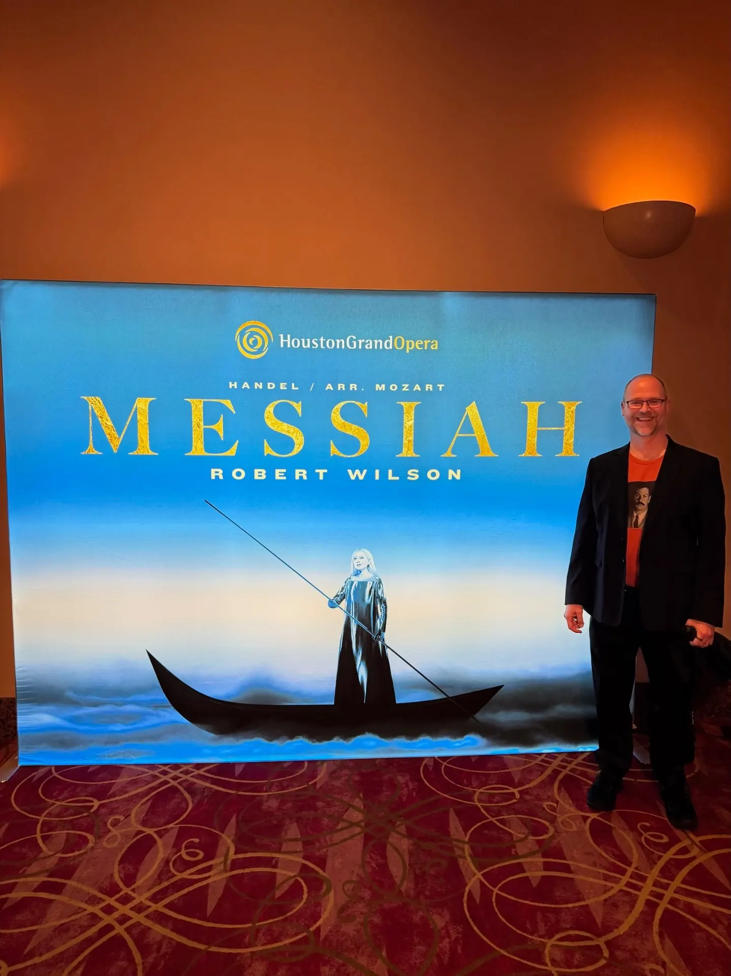 Houston Grand Opera Messiah lobby poster — Handel arranged by Mozart, Robert Wilson production, figure poling a black boat across a pale blue sky, bald man in orange tee and black blazer standing beside it