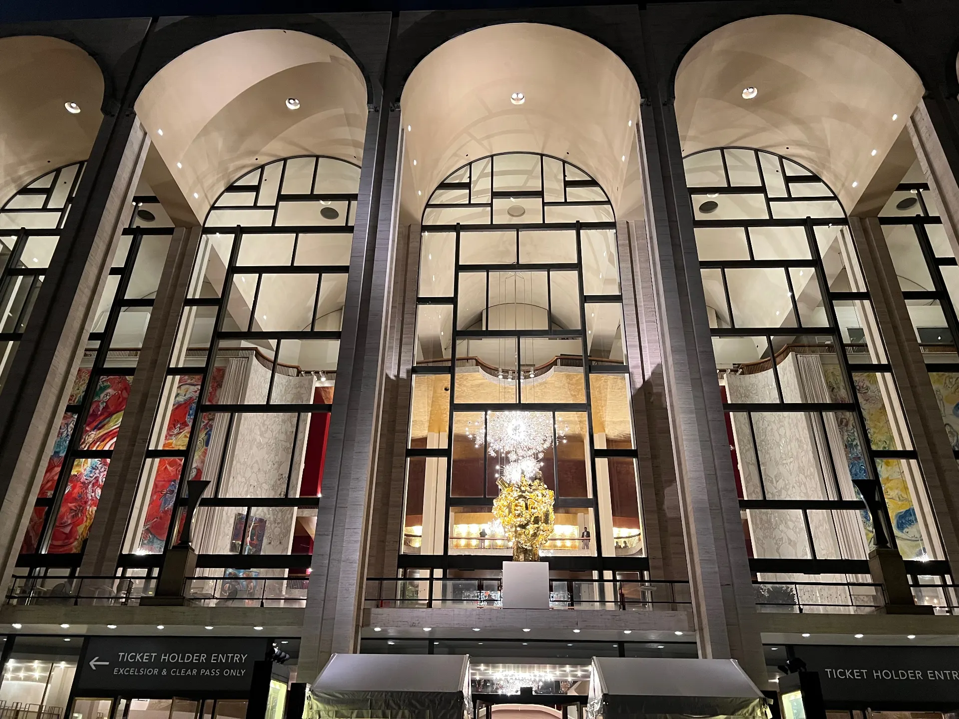 Close-up of the Metropolitan Opera House facade at night, with Chagall murals and chandelier visible through the arched windows