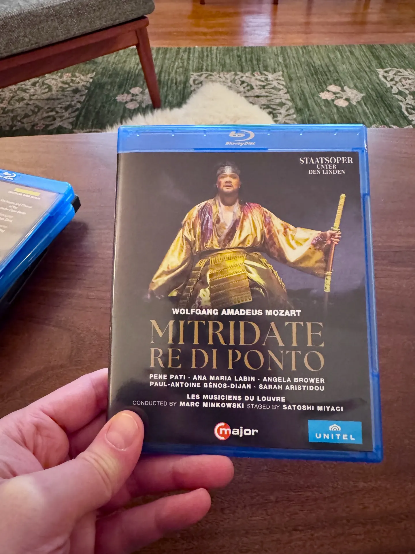 Hand holding the C major Blu-ray of Mozart's Mitridate, re di Ponto from Staatsoper Unter den Linden, Pene Pati on the cover in samurai armor with a katana