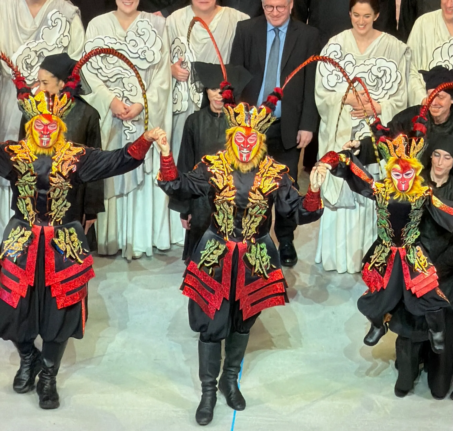 Three performers in glittering red and gold monkey masks with long pheasant-feather plumes bow downstage, robed chorus in cloud-painted white behind them