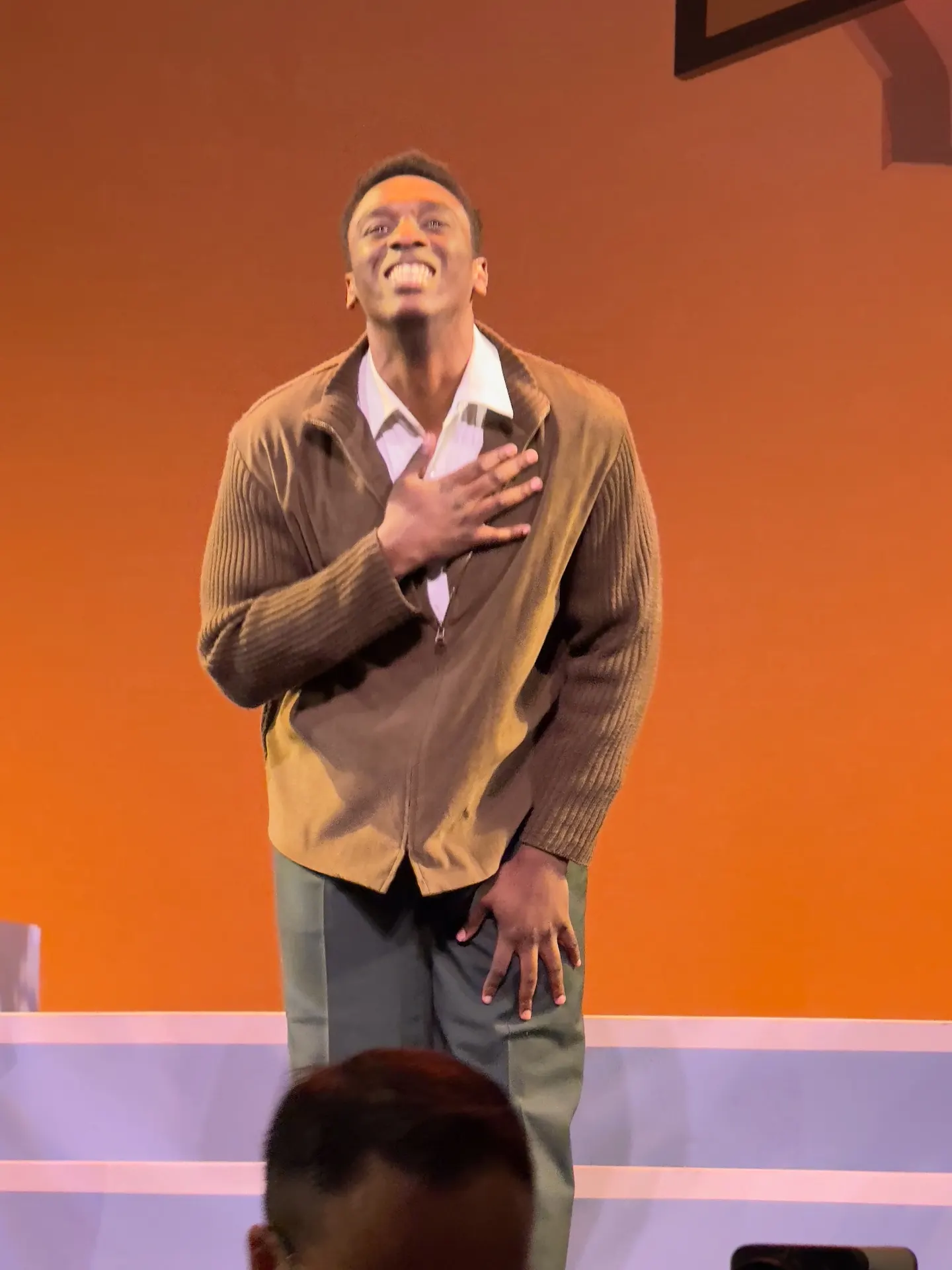 Tenor in a tan zip-up sweater on an orange-walled set, hand to chest at solo bow, colleague in grey suit applauding