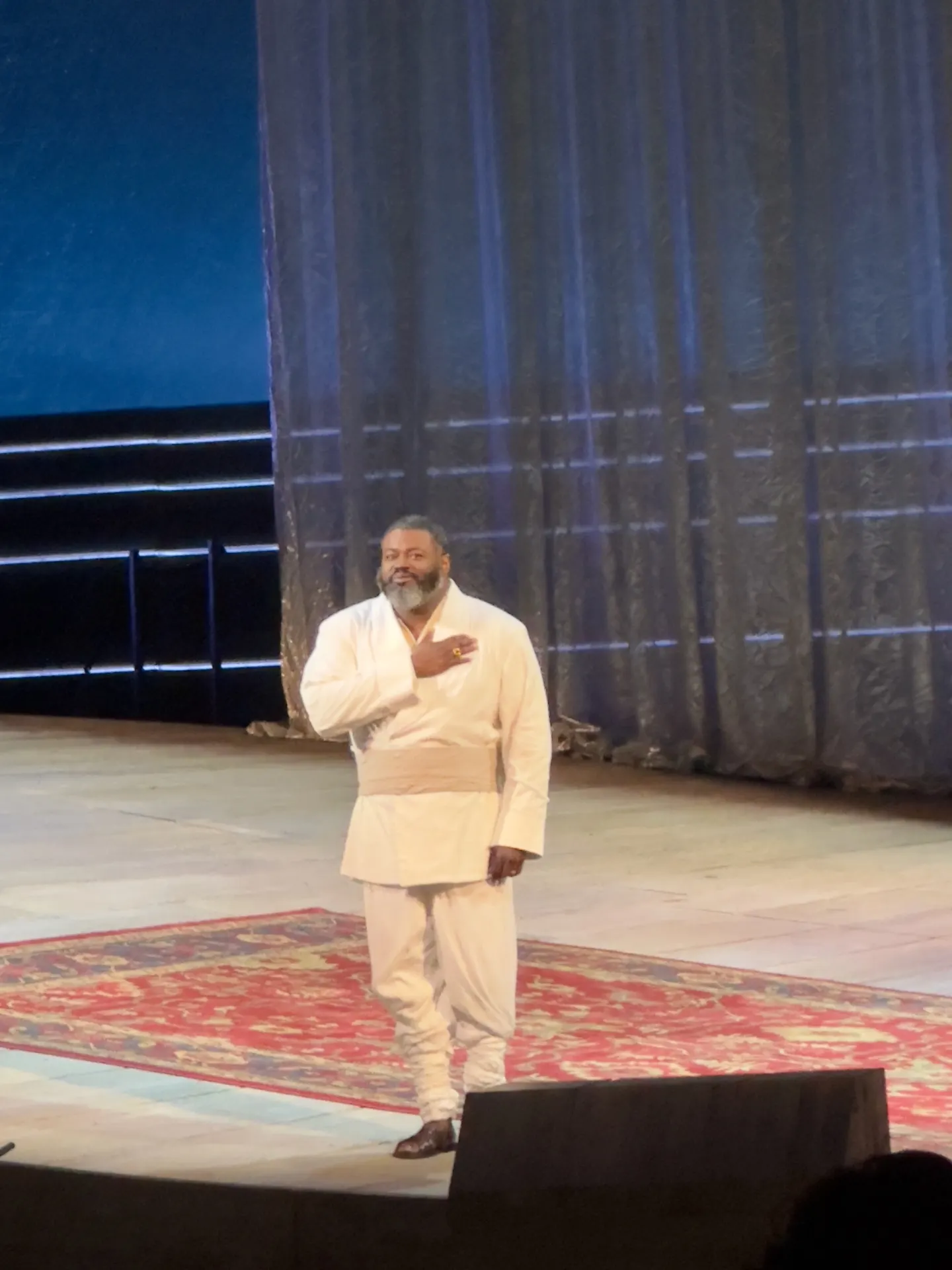Bass-baritone Otello in white robe and sash, alone on a Persian rug at curtain call, hand to chest, gauze curtain behind