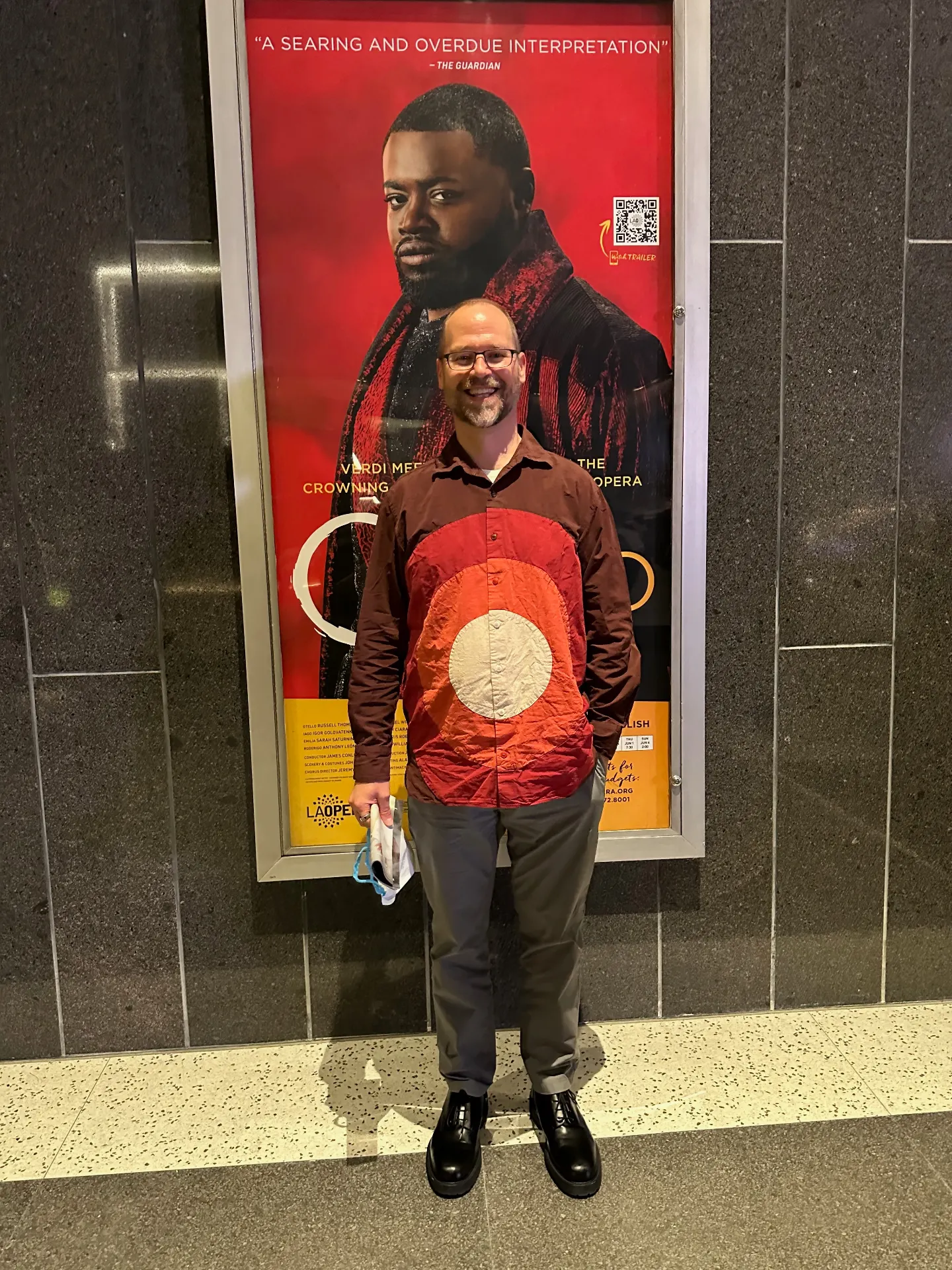 Man in a red-and-rust shirt posing in front of the LA Opera Otello poster, 'A searing and overdue interpretation' quoted from The Guardian