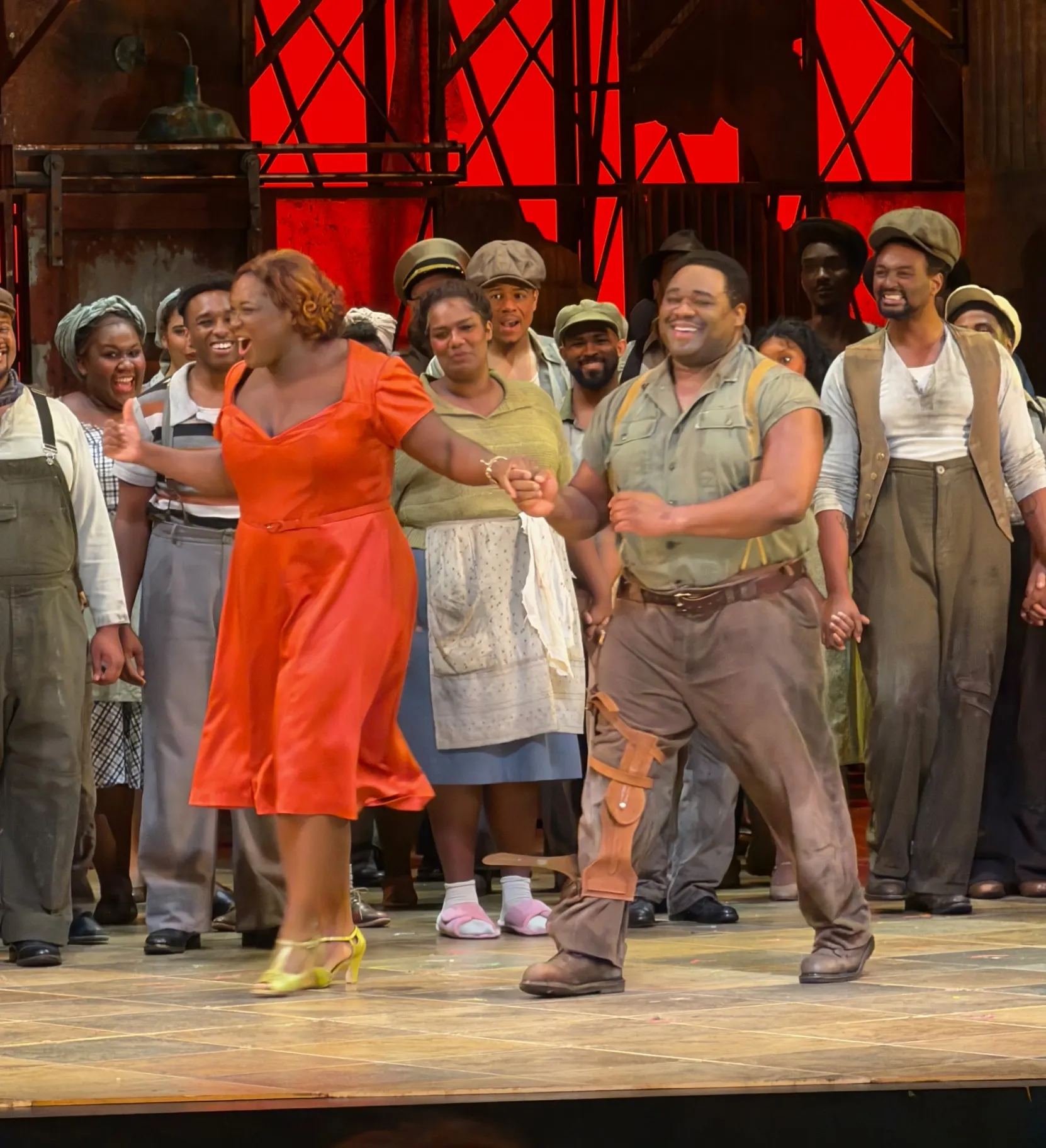 Bess in a red dress and yellow heels striding out for her bow beside Crown in olive shirt and gunbelt, full chorus applauding behind