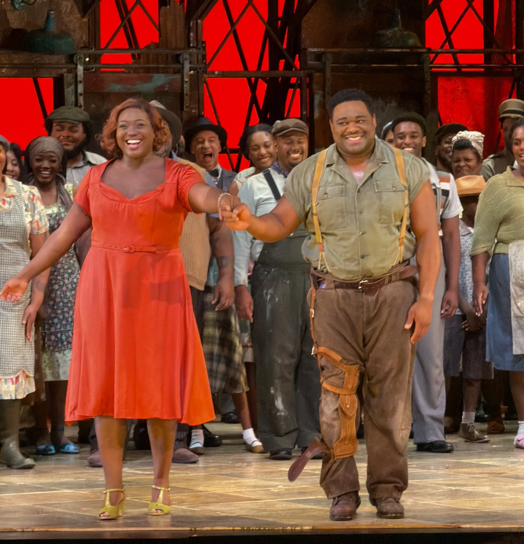 Porgy with his leg brace and Bess in her red dress, hand in hand at the front of the stage, the chorus beaming behind under the red corrugated sky