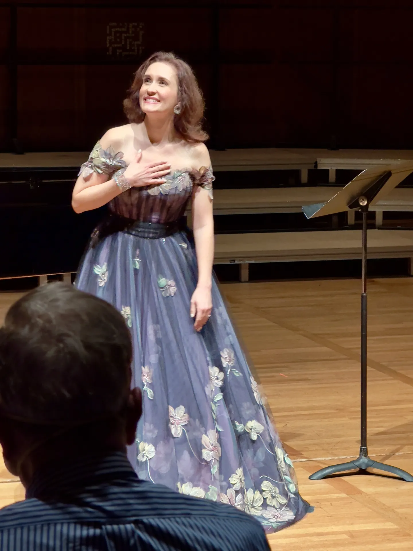 Soprano in a strapless floral-painted gown on a recital stage, hand to chest, smiling out into the hall after a song
