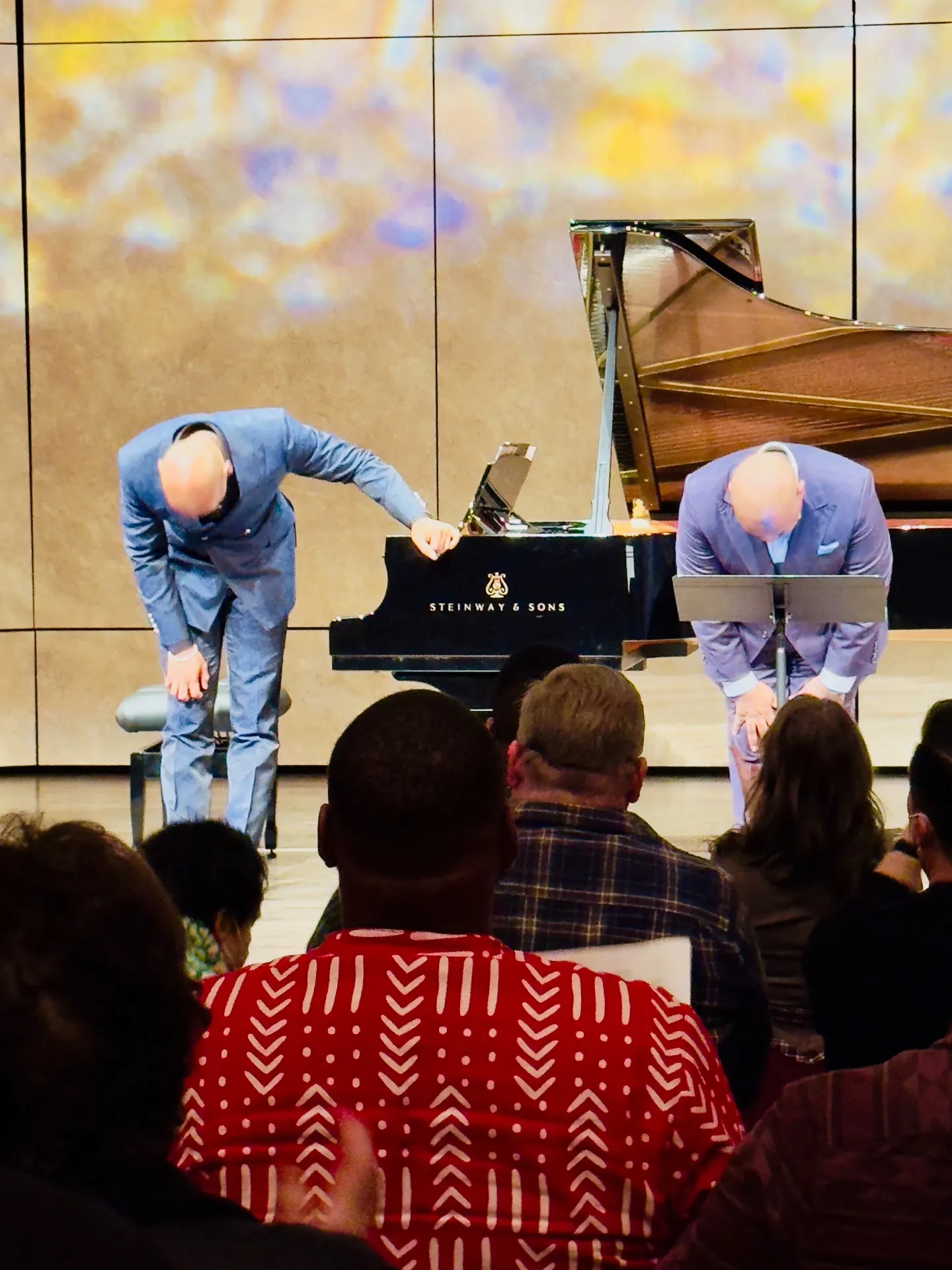 Singer and pianist bowing in matching blue suits beside a Steinway grand, abstract gold-and-purple projection behind them