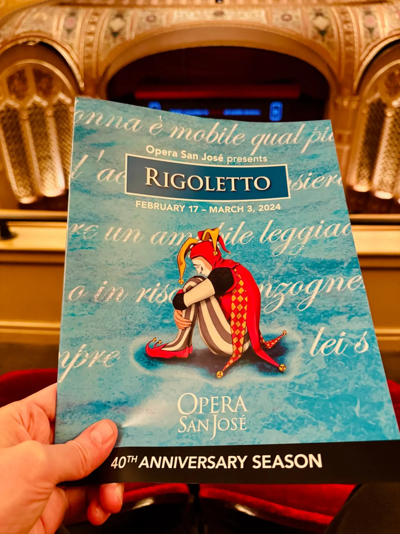 Opera San José Rigoletto program for the 40th Anniversary Season, illustrated jester in red curled up against a pale-blue libretto background, held in the California Theatre