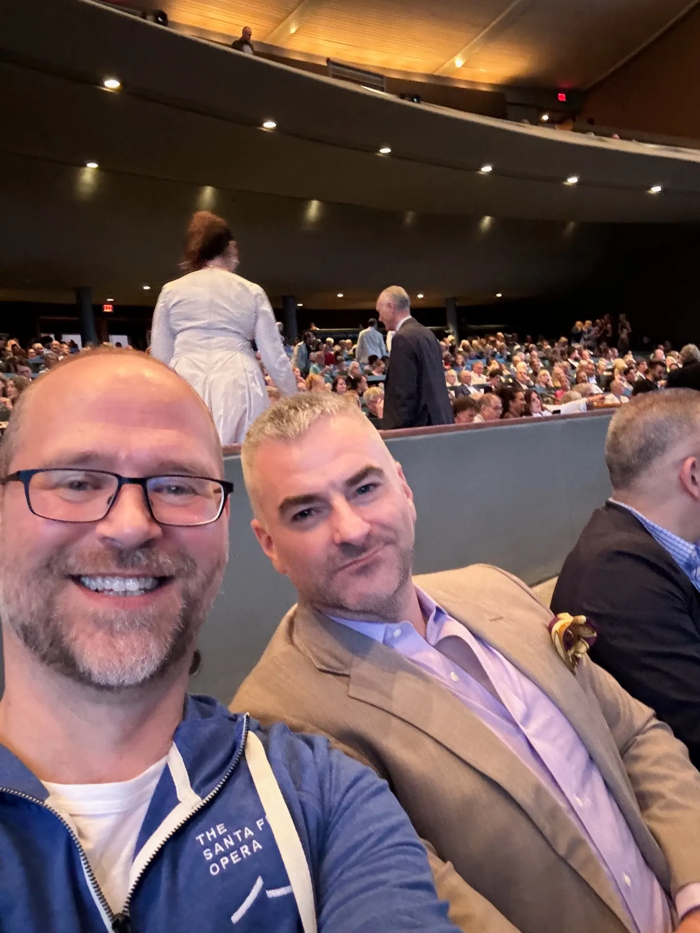 Selfie in a Santa Fe Opera zip-up hoodie with a friend in a tan blazer, audience filing into the open-air house behind
