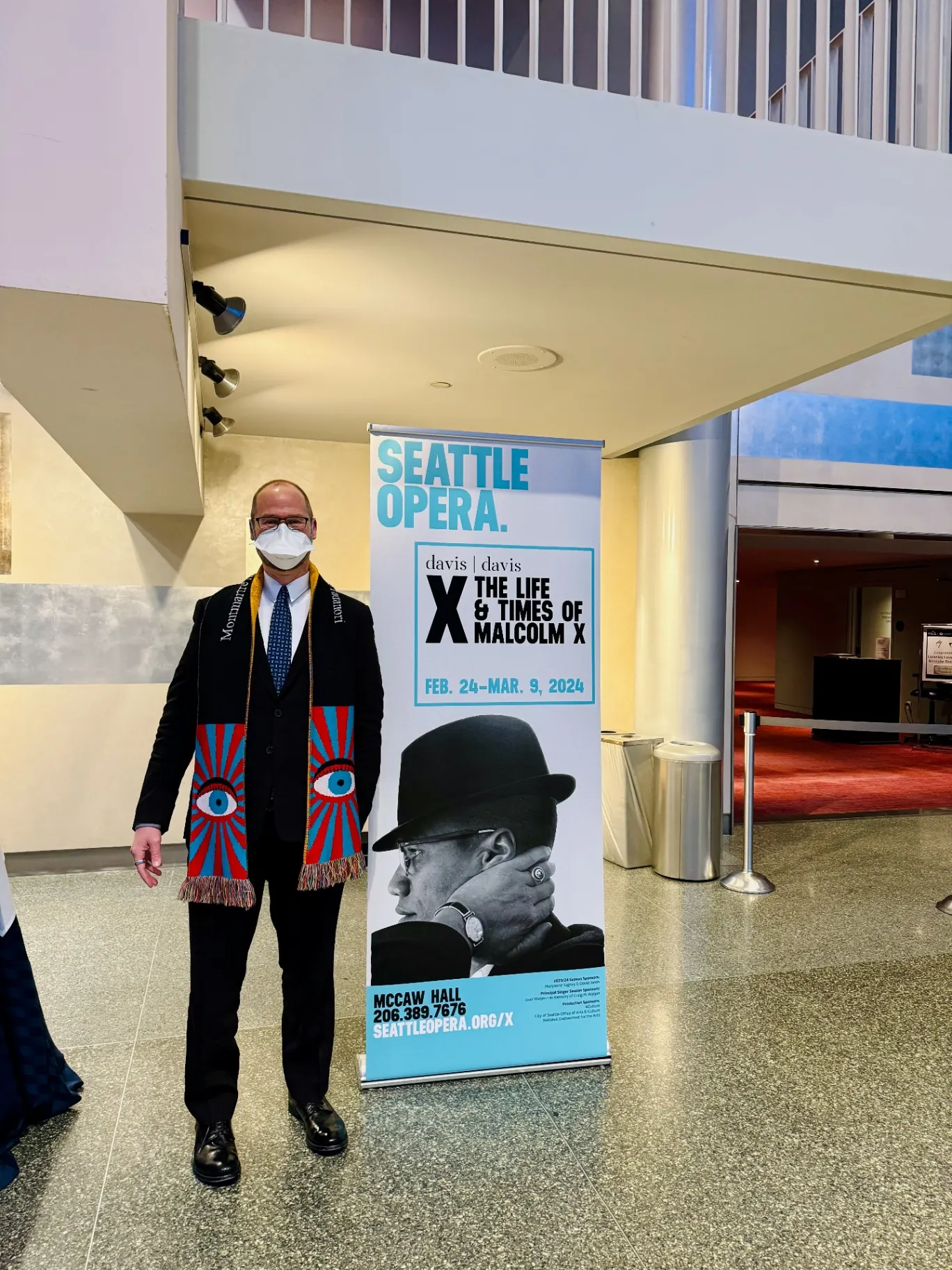 Selfie in N95 mask and an opera-eye scarf next to a Seattle Opera lobby banner for X: The Life and Times of Malcolm X, Feb 24–Mar 9, 2024