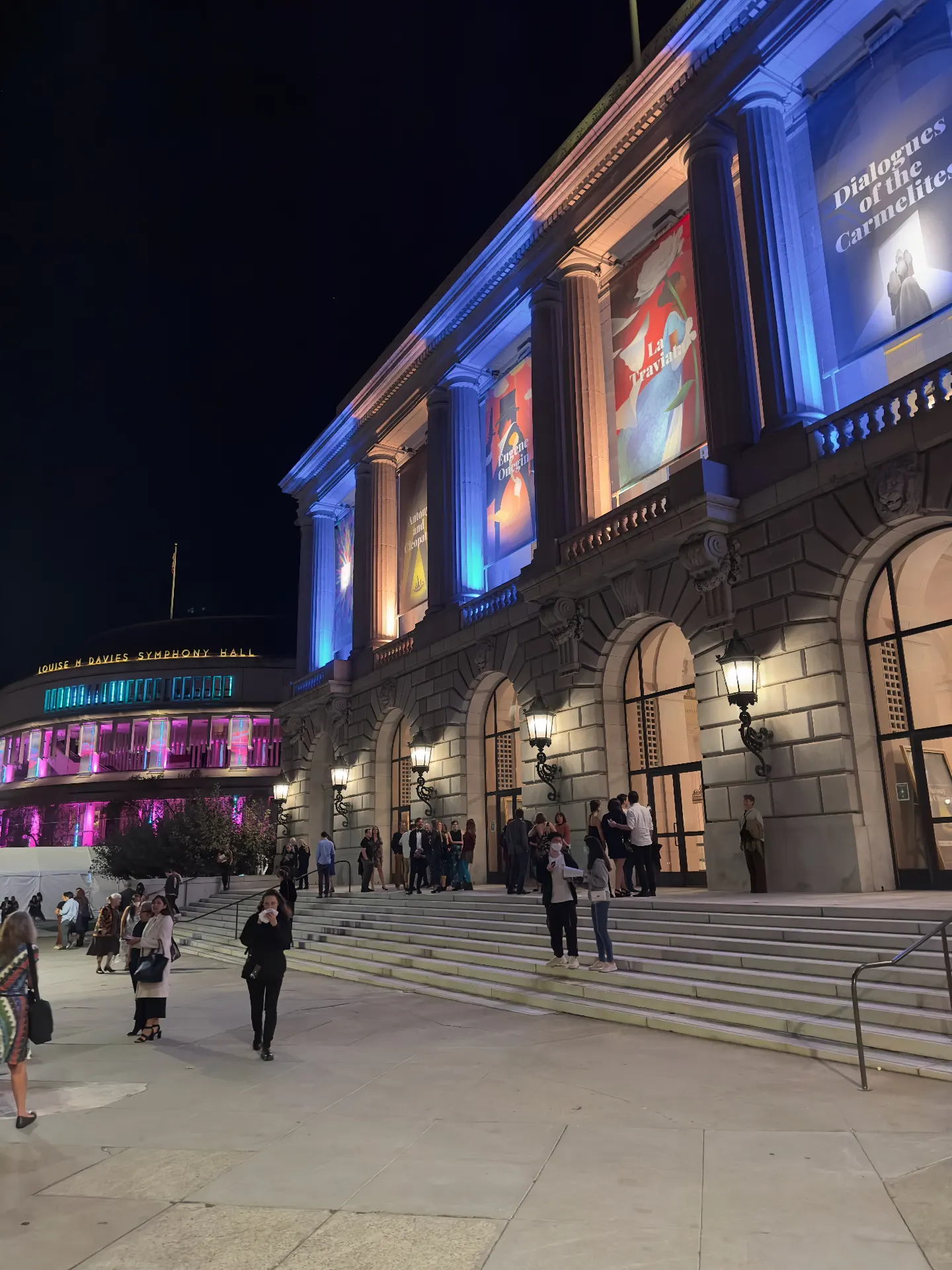 War Memorial Opera House lit blue at night with banners for Dialogues of the Carmelites, La Traviata, and Eugene Onegin, Davies Symphony Hall glowing pink alongside