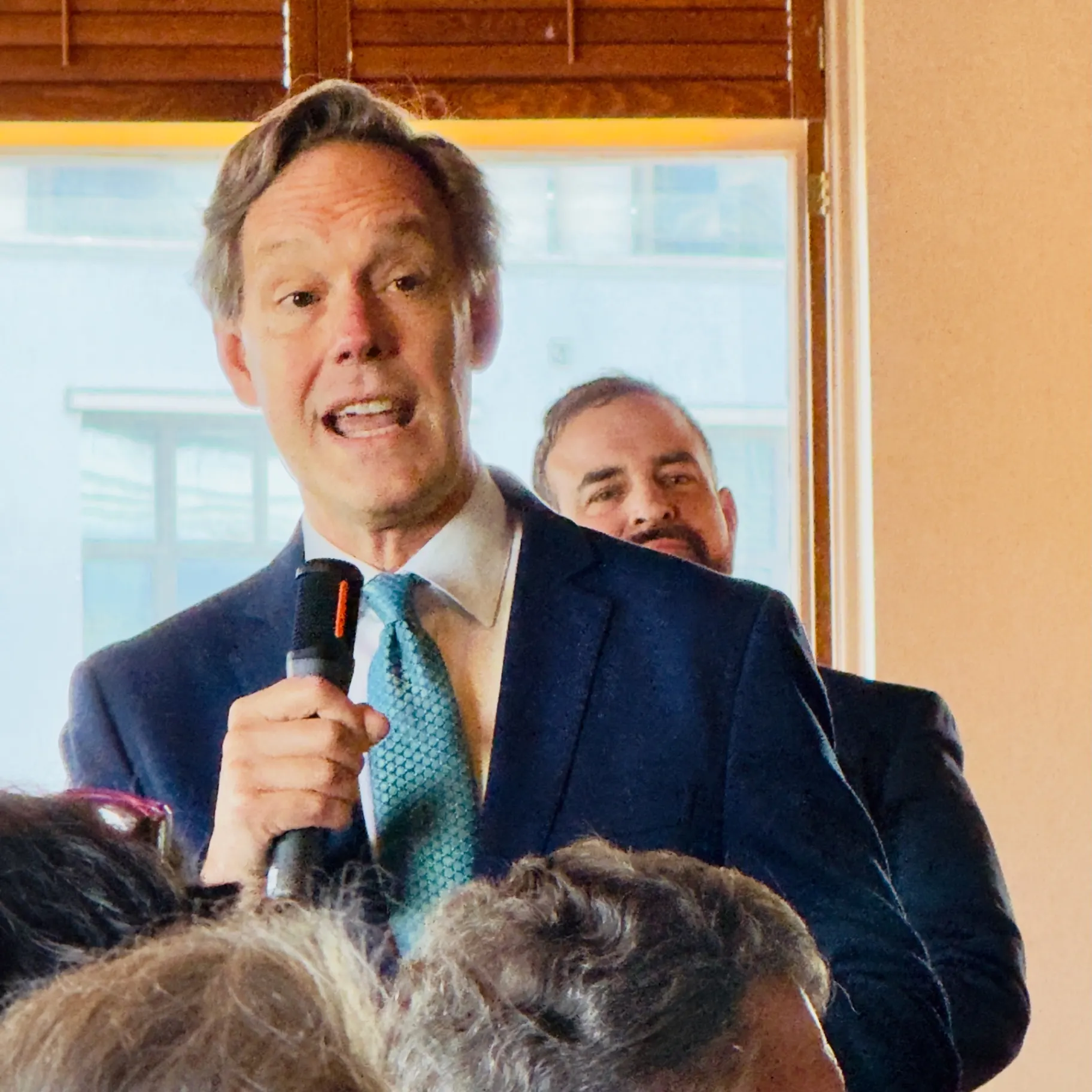 Man in a navy suit and pale blue patterned tie speaking into a handheld microphone at a daytime reception, a colleague listening behind him