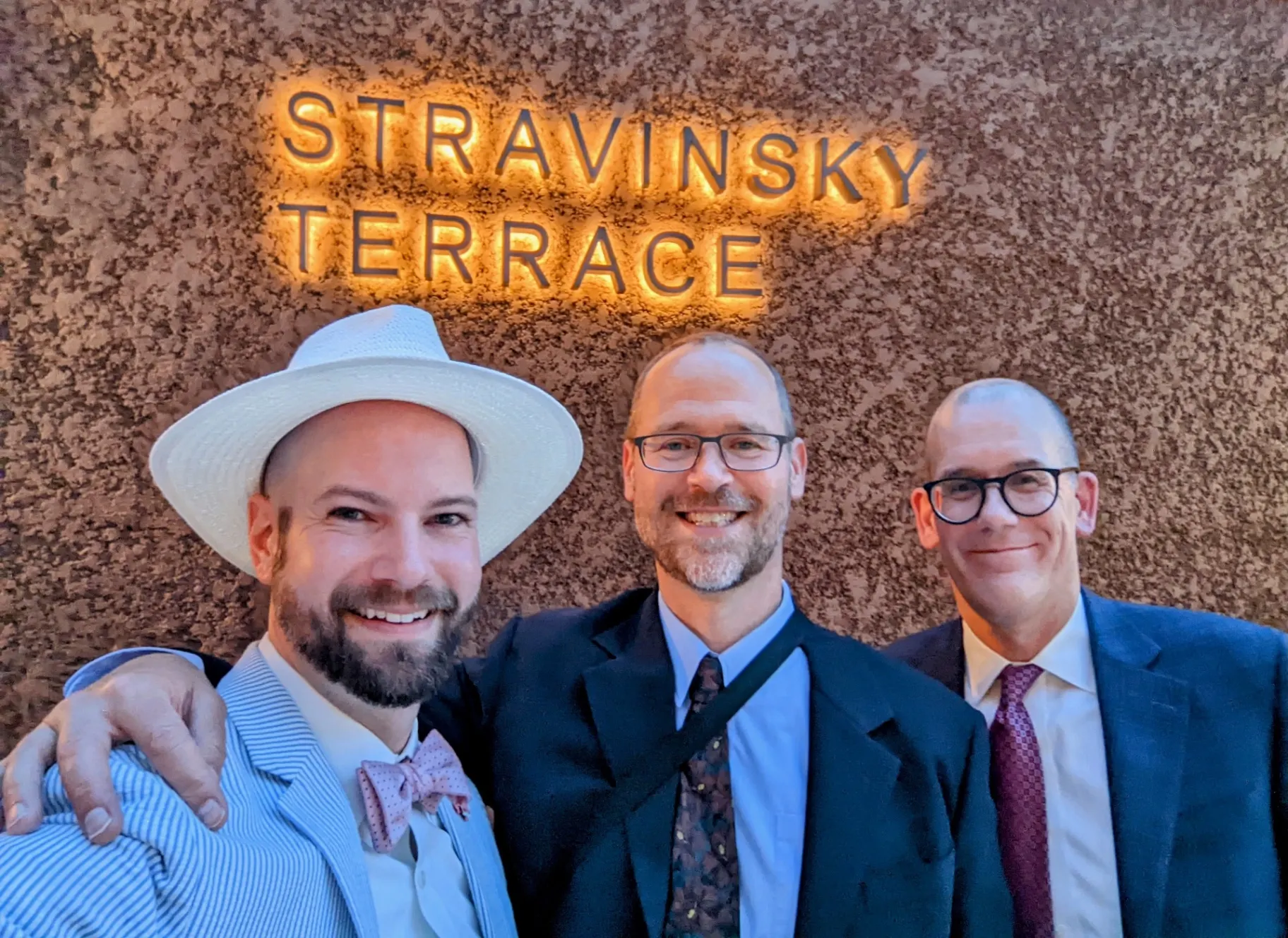 Three friends in jackets and ties posing under the illuminated Stravinsky Terrace sign at the Metropolitan Opera