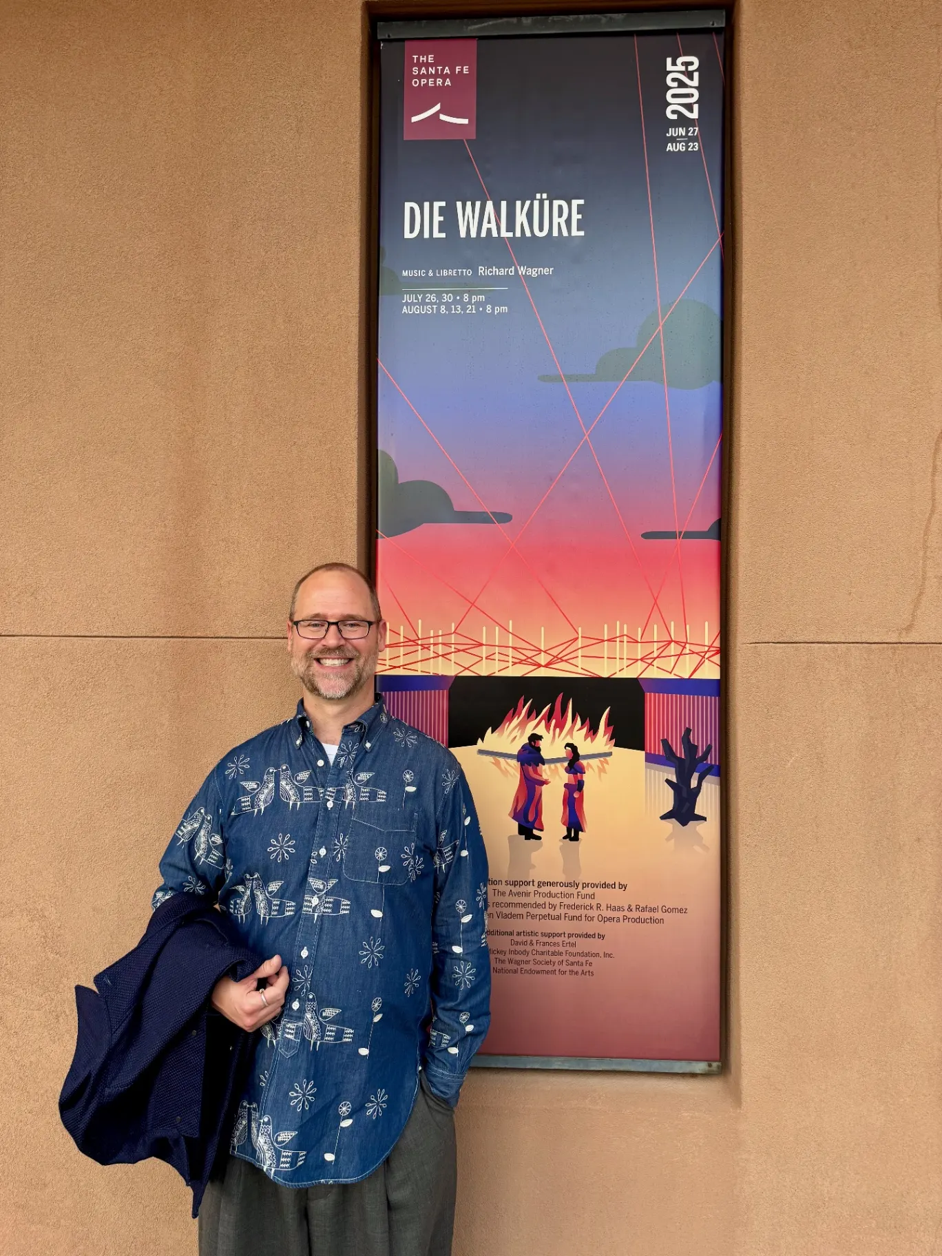 Bald man in a denim shirt printed with white birds, navy blazer over his arm, smiling beside the 2025 Santa Fe Opera Die Walküre poster mounted on adobe wall