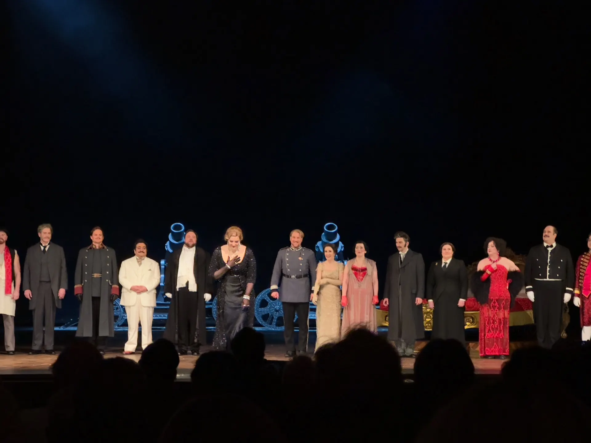 Wide curtain call line at the Metropolitan Opera with cannons and gun carriages upstage, full principal cast bowing in tsarist-era costume