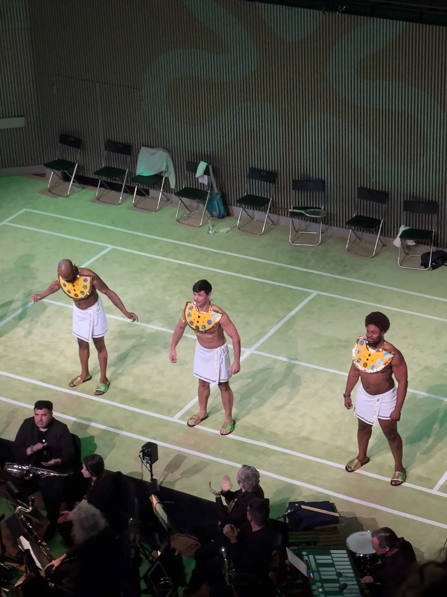 Three singers in white kilts and gilded Egyptian collars standing on a green tennis-court set, orchestra visible below